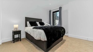 Un dormitorio moderno con cabecera oscura, cama con ropa de cama blanca y negra, mesita de noche con lámpara y vista al exterior a través de una ventana.
