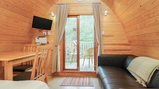 Een gezellig interieur van een blokhut met een houten plafond, een aan de muur bevestigde televisie, een eettafel met stoelen, een bank en een glazen schuifdeur die leidt naar een terras.
