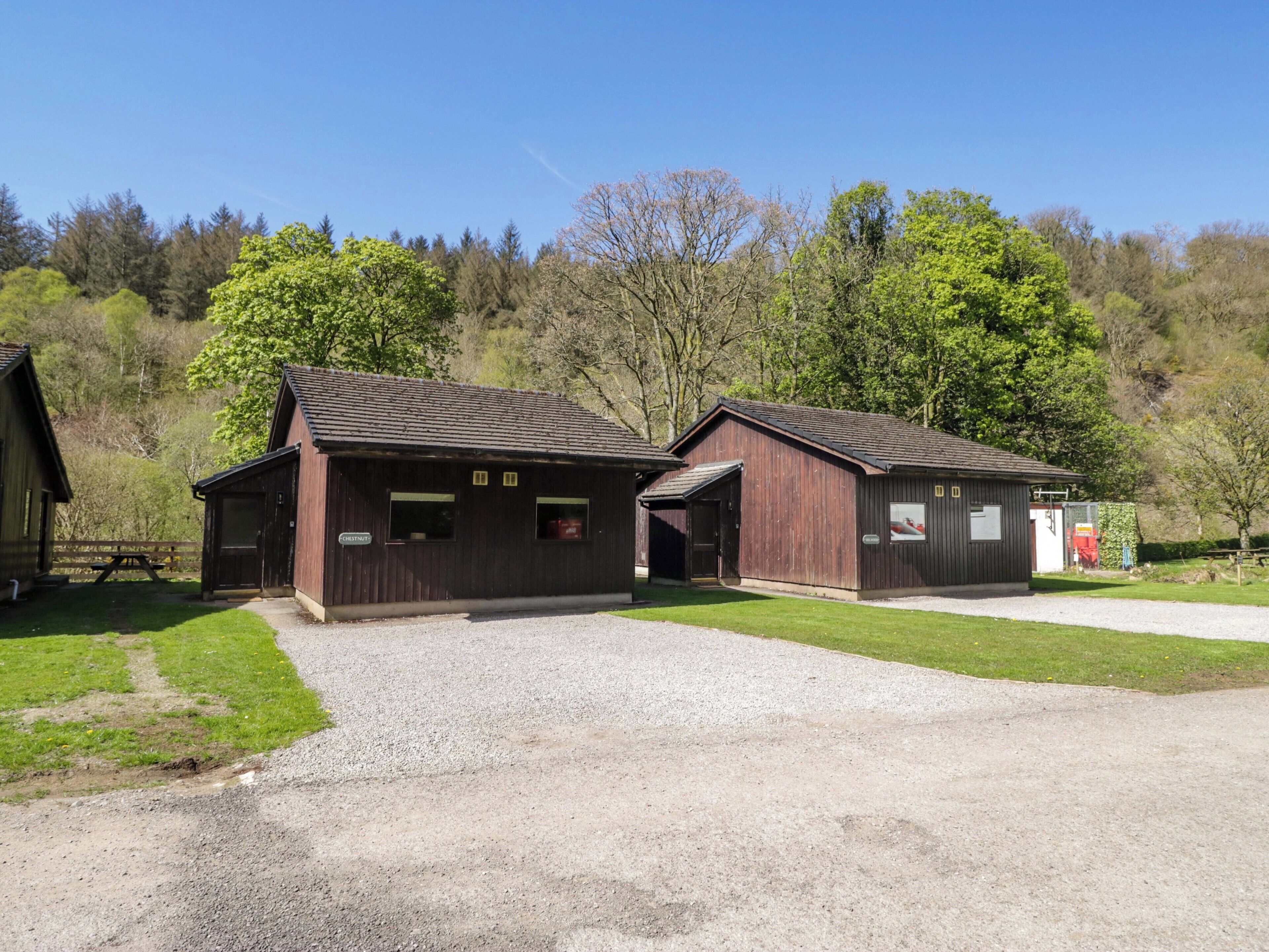 Two wooden cabins with a gravel driveway and picnic table, surrounded by trees and a clear sky.