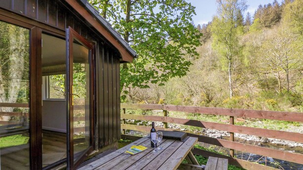 A wooden deck with a glass door leading to a room, a picnic table, and a view of a river and forest. - Chestnut Timber Lodge (Keswick)