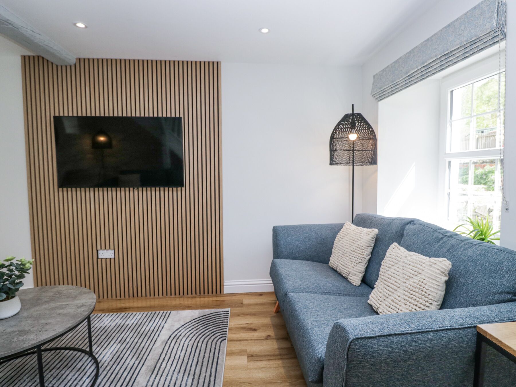 Een moderne woonkamer met een grijze bank, een hangende televisie, een vloerlamp en een wandpaneel van hout.