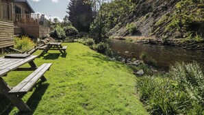 Een picknickplaats aan de rivier met houten banken en een picknicktafel, omgeven door weelderige begroeiing en een houten hut.