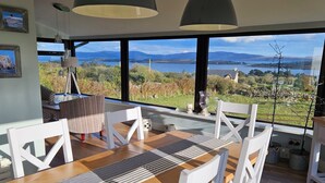 A dining area with a wooden table and white chairs, large windows offering a view of a lake and mountains. - Casey House (Castletownbere)