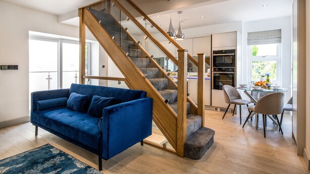 A modern living room with a blue sofa, a wooden staircase, and a dining area with a table and chairs. - The Old Sweet Shop (Conwy)