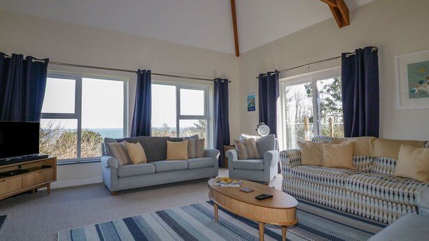 A spacious living room with two sofas, a coffee table, and a TV. Large windows with curtains offer views of the outdoors.