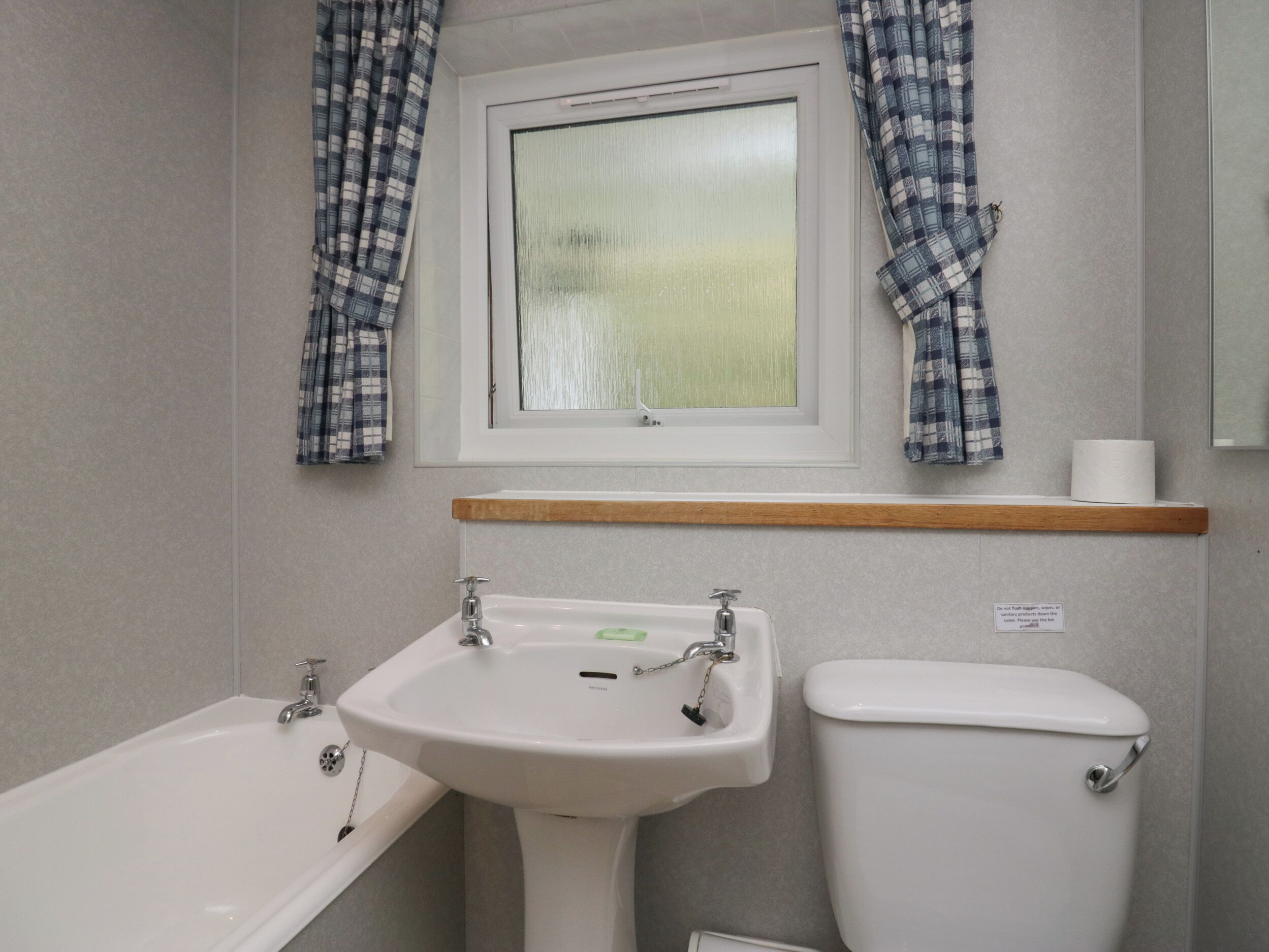 A bathroom with a white sink, a toilet, and a bathtub. There is a window with curtains and a wooden shelf above the sink.
