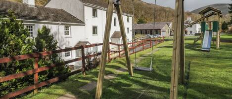 Un parque infantil con columpios y un tobogán, rodeado por una valla de madera, con casas al fondo.