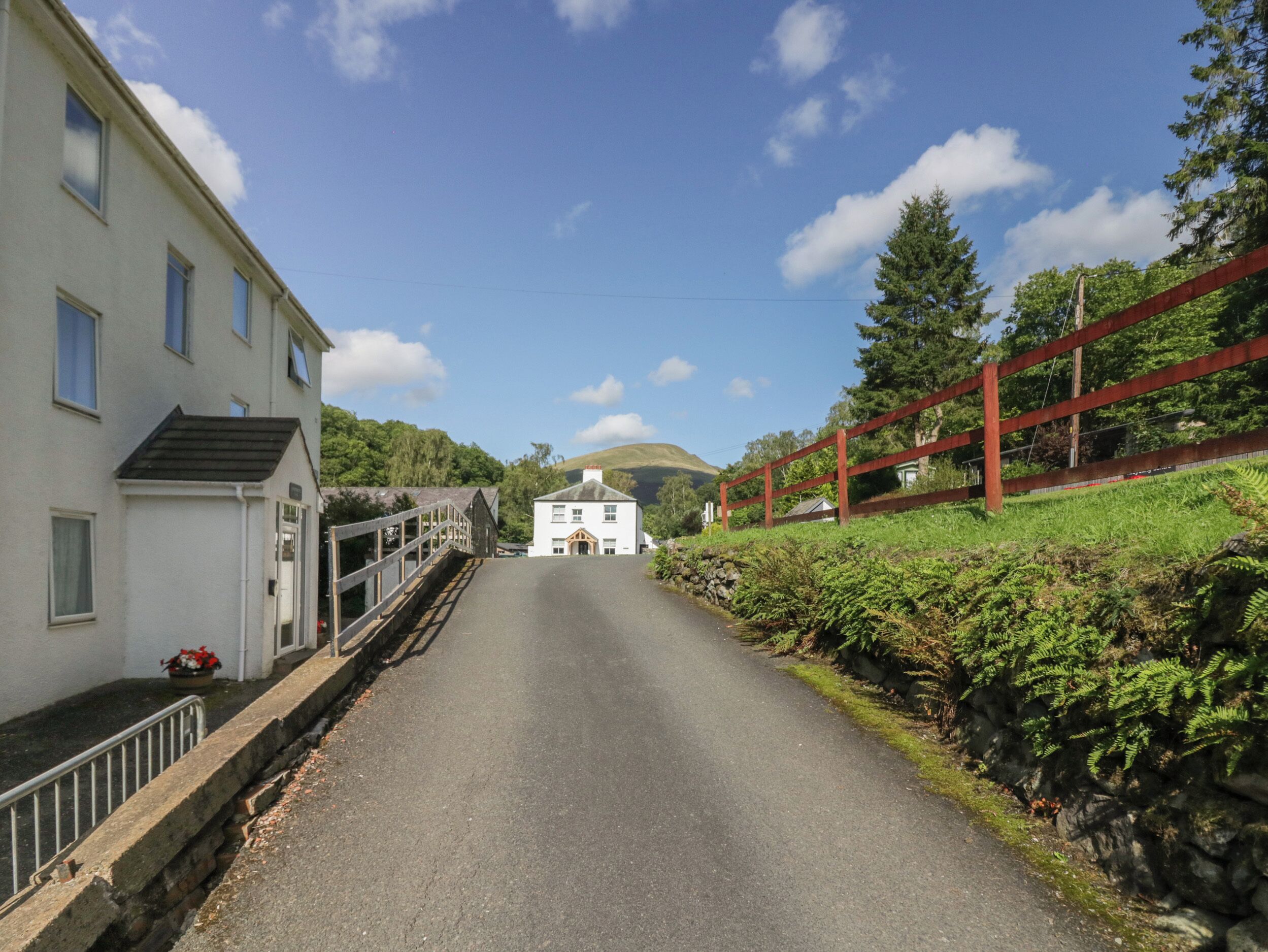 A narrow road flanked by buildings and greenery, leading to a white building with a gabled roof.