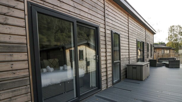 A wooden cabin with large sliding glass doors, a deck with outdoor furniture, and a small attached structure with a window. - The Bothy (Keswick)