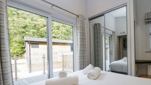 A bedroom with a large bed, a view of a wooden deck and a small building, and a sliding glass door leading to a balcony. - The Bothy (Keswick)
