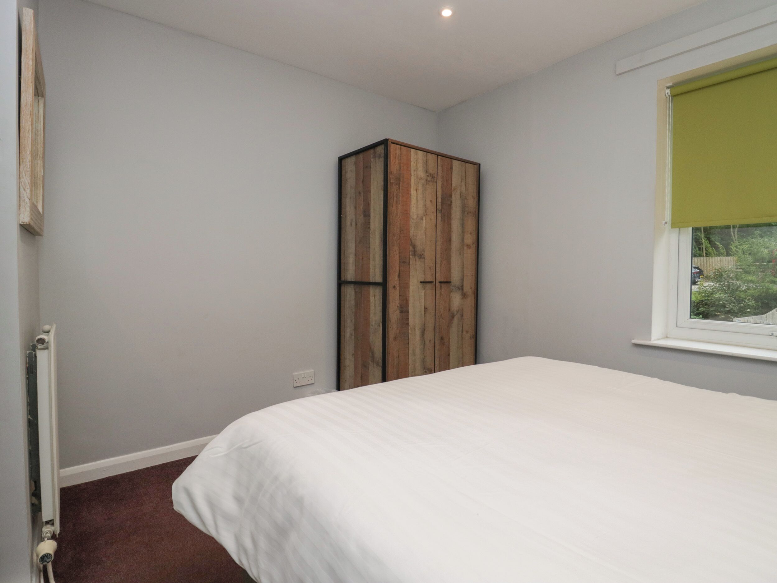 A bedroom with a bed, a wooden wardrobe, a radiator, and a window with a green blind.