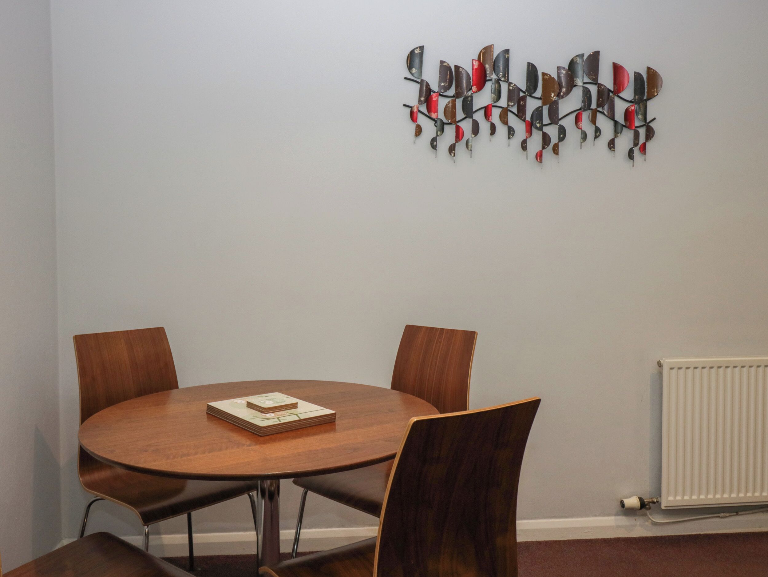 Une table ronde en bois, accompagnée de quatre chaises, un élément décoratif en métal fixé au mur et un radiateur blanc.