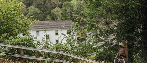 A white building with a sloped roof, surrounded by dense green trees and a wooden bridge.