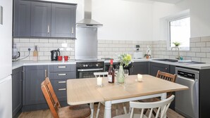Une cuisine moderne avec des armoires grises, une hotte de cuisinière en acier inoxydable, un four encastré et un lave-vaisselle. On y trouve une table à manger en bois avec des chaises, ainsi qu’une fenêtre qui laisse entrer la lumière naturelle.
