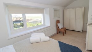 A bedroom with a large window, a bed with white linens, a wicker chair, and a wardrobe. - Little Haven (Bamburgh)
