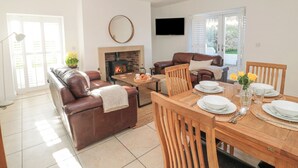 A living room with a fireplace, a brown leather sofa, a wooden dining table set for four, and a television mounted on the wall. - Little Haven (Bamburgh)