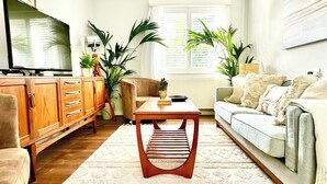 A living room with a wooden TV stand, a sofa, a coffee table, and several potted plants. - The Shingles (Sandown)