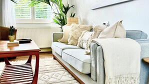 A living room with a grey sofa, a wooden coffee table, a potted plant, and a large window with blinds. - The Shingles (Sandown)