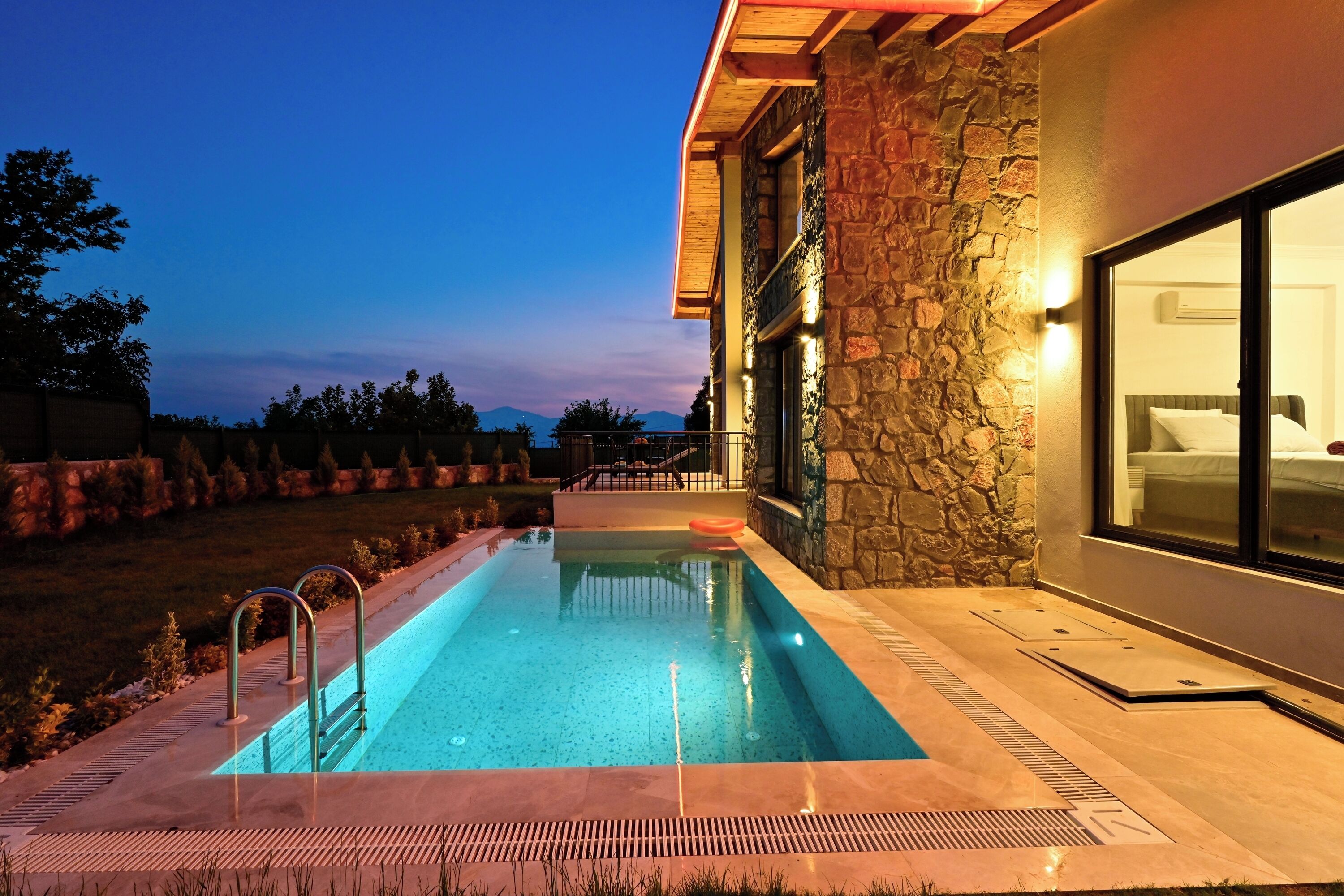 A swimming pool adjacent to a stone building with large windows, a well-lit exterior, and a view of the mountains in the background.