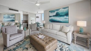 A living room with a large wave painting, a ceiling fan, and a coffee table with magazines. - SW Facing Beachfront Condo | Sweeping Ocean Views (Marco Island)