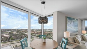 A modern living room with a large window, a round wooden table, and a sofa. - SW Facing Beachfront Condo | Sweeping Ocean Views (Marco Island)