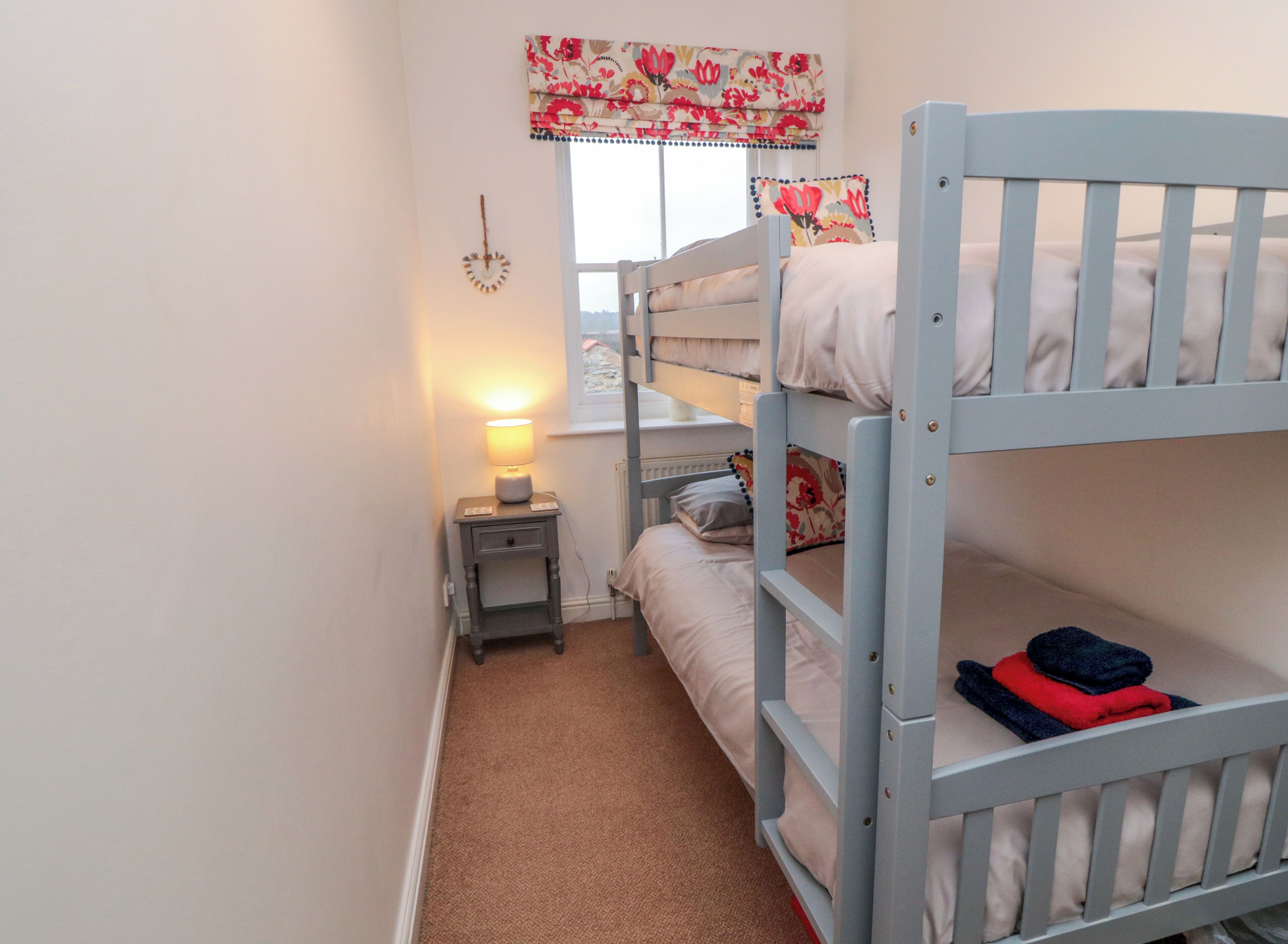 Une chambre avec un lit superposé, une petite table de chevet, une lampe et une fenêtre ornée de rideaux à motifs floraux.