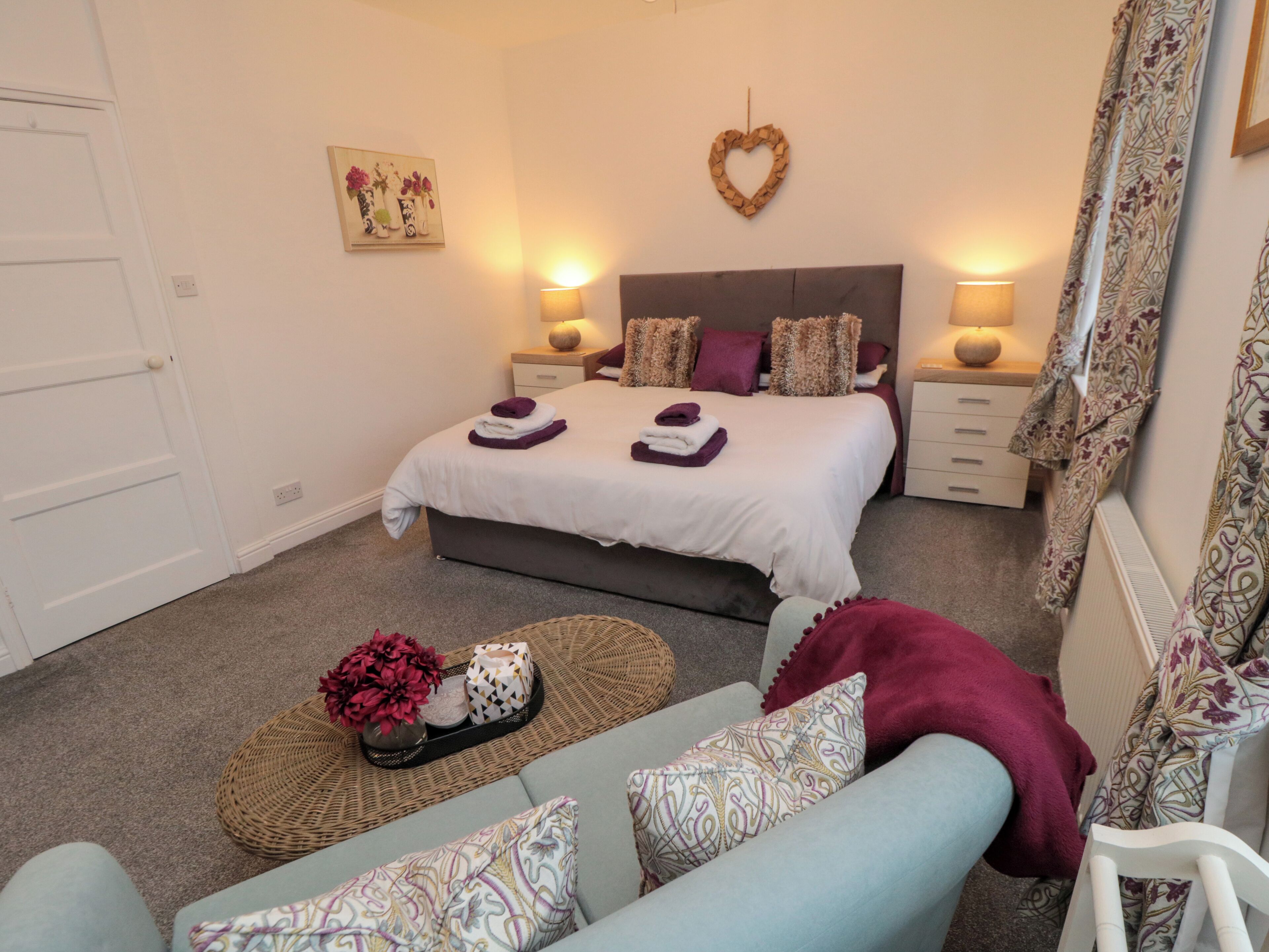A bedroom with a bed, two bedside tables, a nightstand, a sofa, a decorative basket, and a heart-shaped wall decoration.