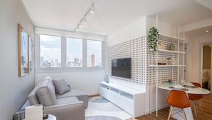 A modern living room with a grey sofa, a white TV stand, a dining table with orange chairs, and a city view through large windows. - Extension Berrini (Sao Paulo)