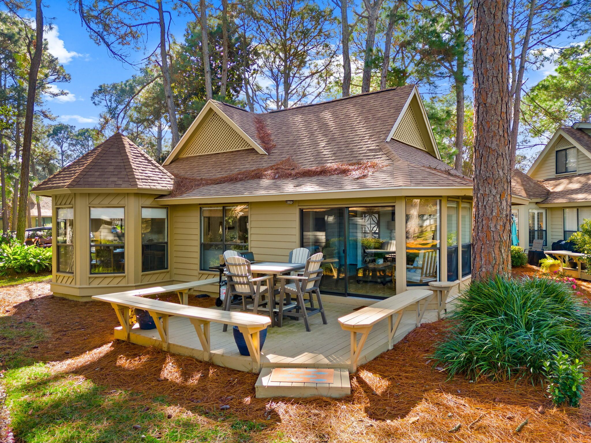A two-story house with a covered patio, a dining area with chairs and a table, and a small garden area with plants and a bench.