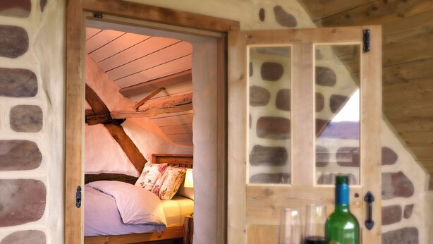 A cozy bedroom with a stone wall, wooden ceiling beams, and a bed with floral pillows.