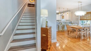Eine Treppe führt zu einem Essbereich mit Holztisch und -stühlen, im Hintergrund ist eine Küche zu sehen und auf einer Kommode steht eine dekorative Lampe.