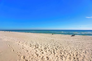 A wide sandy beach with footprints, a clear blue sky, and calm ocean waves.
