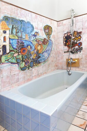A bathroom with a large bathtub, colorful tile mural, and a showerhead.