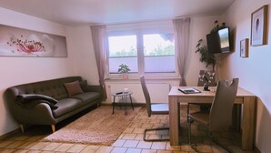 Un salon confortable avec un canapé, une petite table, un fauteuil, un téléviseur et une fenêtre avec des rideaux.