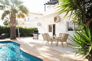 Un espace aménagé au bord de la piscine, avec un barbecue, des sièges d’extérieur et un palmier.