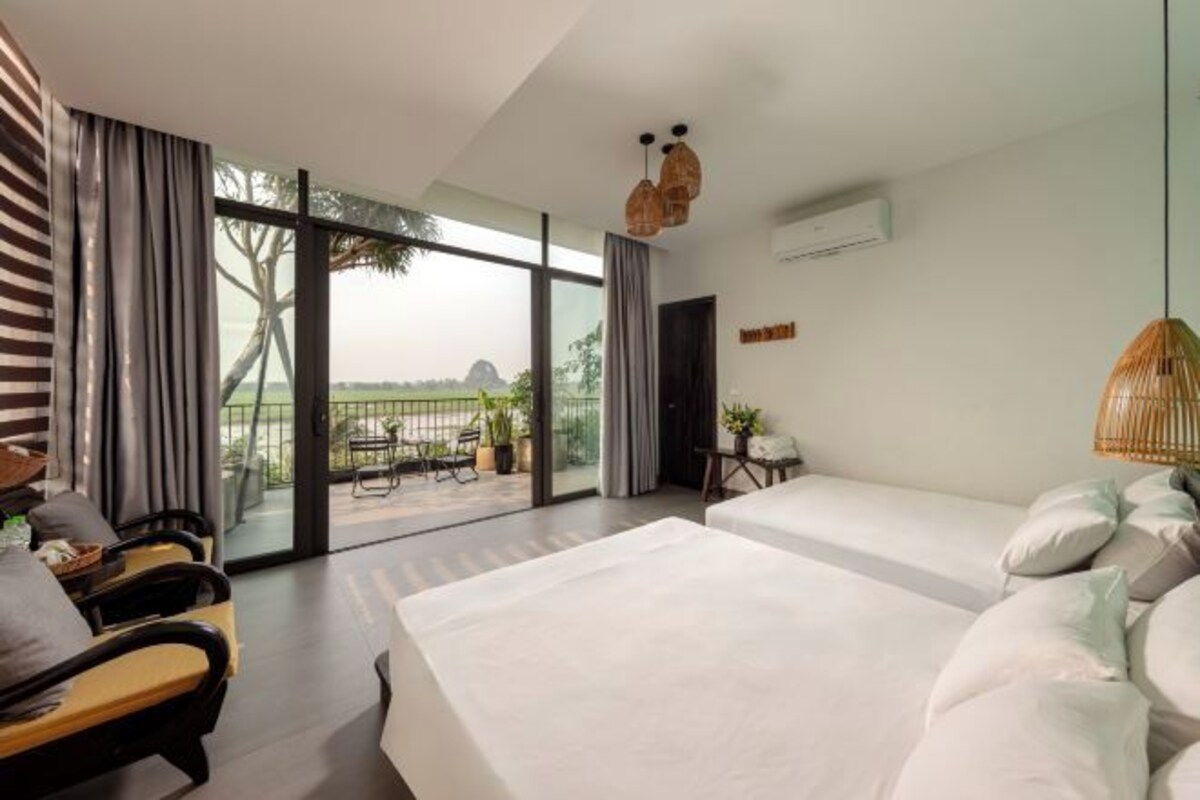 A modern hotel room with a large sliding glass door leading to a balcony, two single beds, a sofa, and a small table.