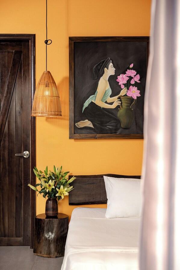 A bedroom with a wooden door, a hanging lamp, a painting of a woman holding flowers, and a bedside table with a vase of flowers.