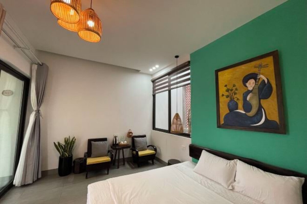 A hotel room with a large bed, a green accent wall, a painting, a window with blinds, a small table with a plant, and a chair.