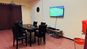 A dining area with a table, chairs, a TV mounted on the wall, and a clock. - Adrenaline Lodge and Tours (Livingstone)