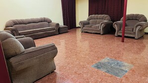 A living room with a brown sofa set, a smaller armchair, and a coffee table. The room has a tiled floor and a red pillar. - Adrenaline Lodge and Tours (Livingstone)