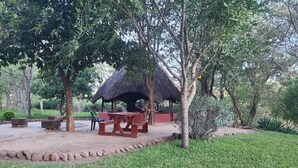 A thatched-roof pavilion with picnic tables and benches in a grassy area surrounded by trees. - Adrenaline Lodge and Tours (Livingstone)