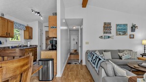 Een moderne keuken met houten kastjes, een eethoek met tafel en stoelen, een grijze bank en een gang naar een slaapkamer.