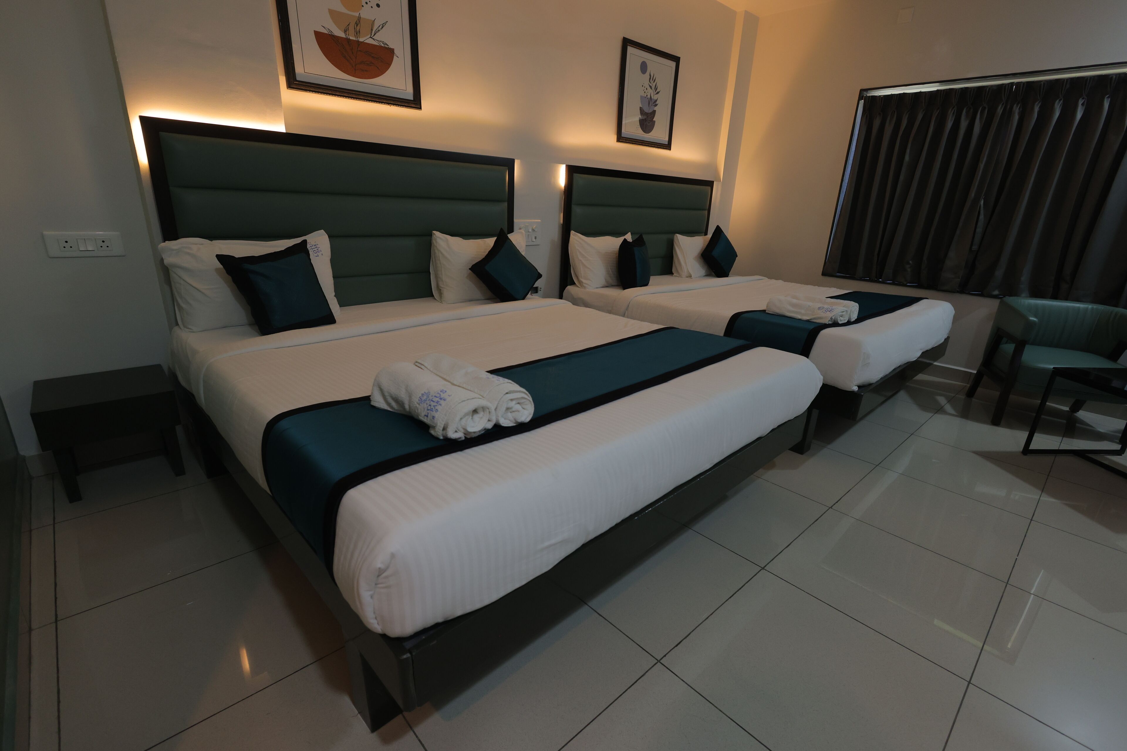 Habitación de hotel con dos camas, cada una con cabecera, ropa de cama blanca y una toalla enrollada sobre la cama. Se observa una silla y una ventana con cortinas.