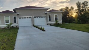 A two-story house with a central garage, surrounded by a well-maintained lawn and trees. - Modern Place,  3-bedroom house with WiFi, AC, spacious in welcoming Lehigh Acres (Lehigh Acres)