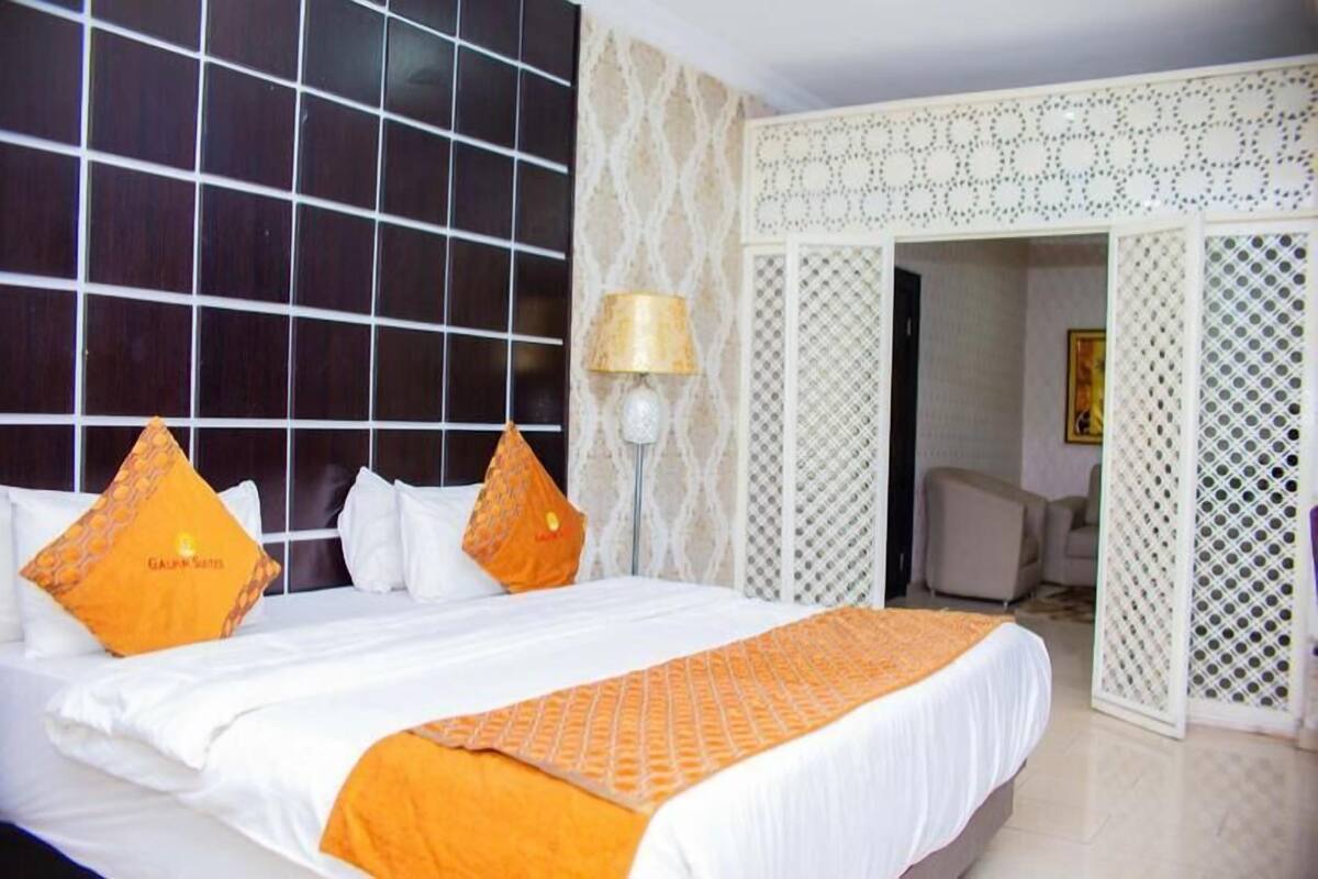 A hotel room with a large bed, white bedding, and orange accents. There is a lamp on the side table and a decorative screen in the doorway.