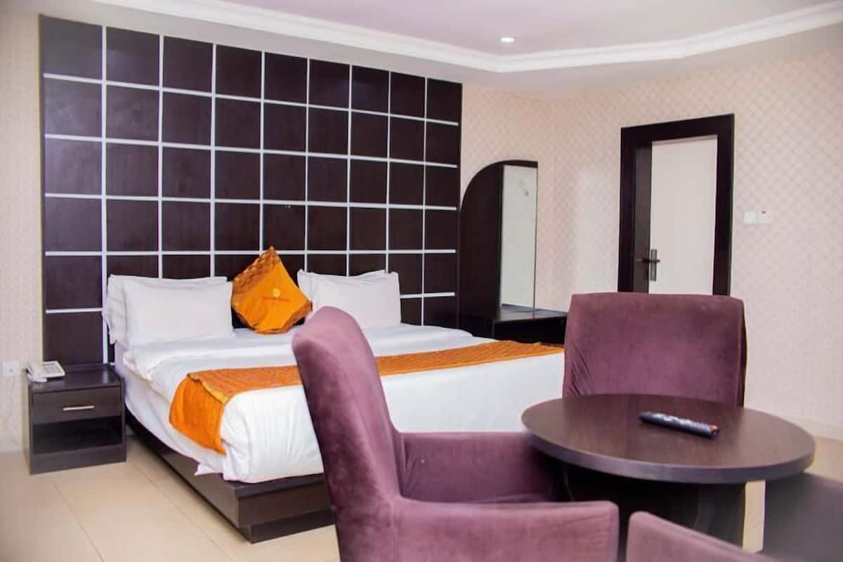 A hotel room with a large bed, a bedside table, a chair, and a small round table with a remote control.