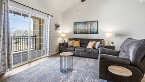 Uma sala de estar com sofá, duas mesinhas de cabeceira com abajures, um ventilador de teto e uma grande janela com cortinas.