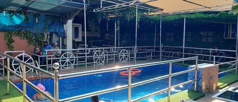 Una piscina exterior con valla metálica, rodeada por un área techada con sillas y mesas.