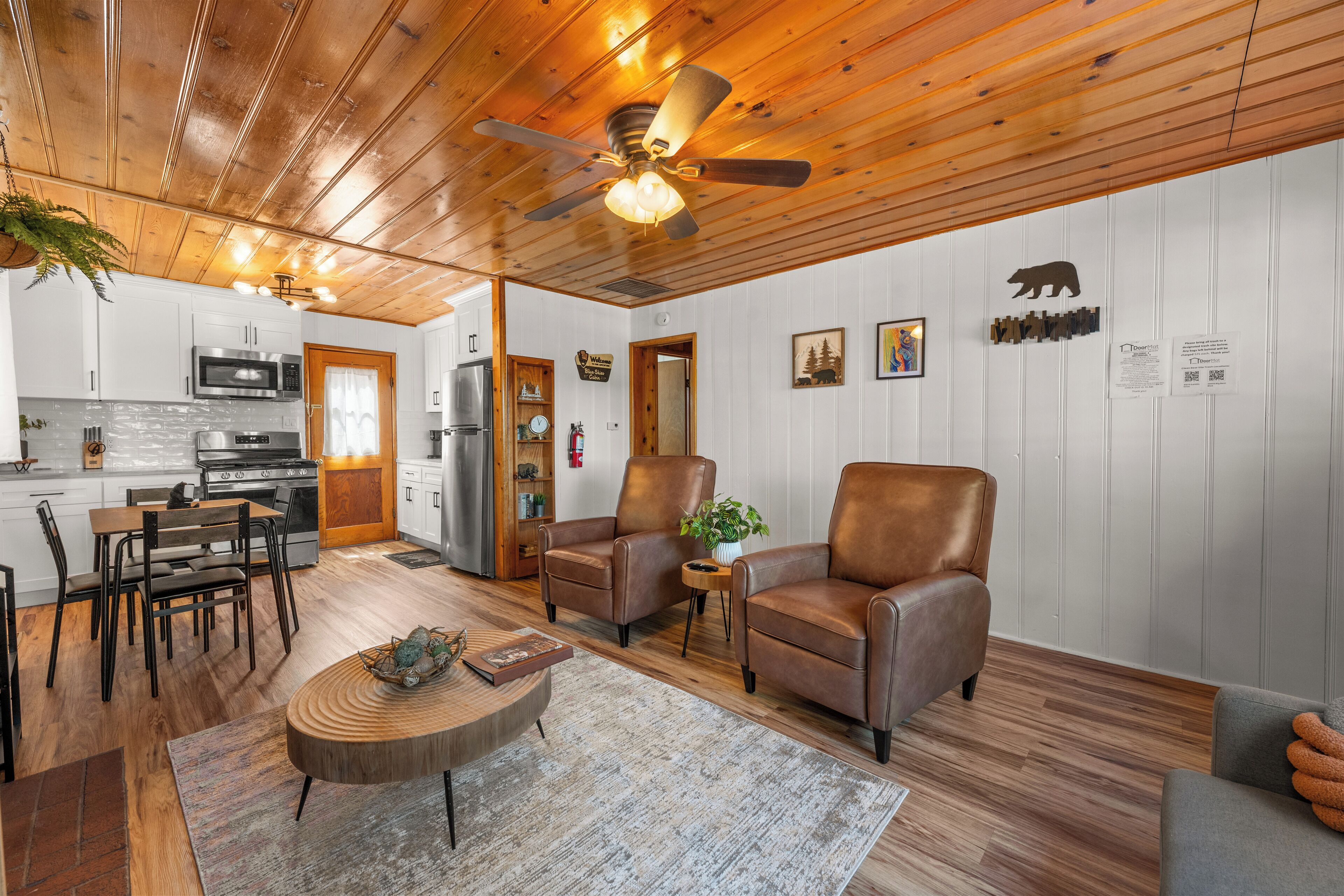 A modern kitchen with white cabinets, a microwave, stove, and a ceiling fan. There are two leather chairs, a coffee table, and a rug in the living area.