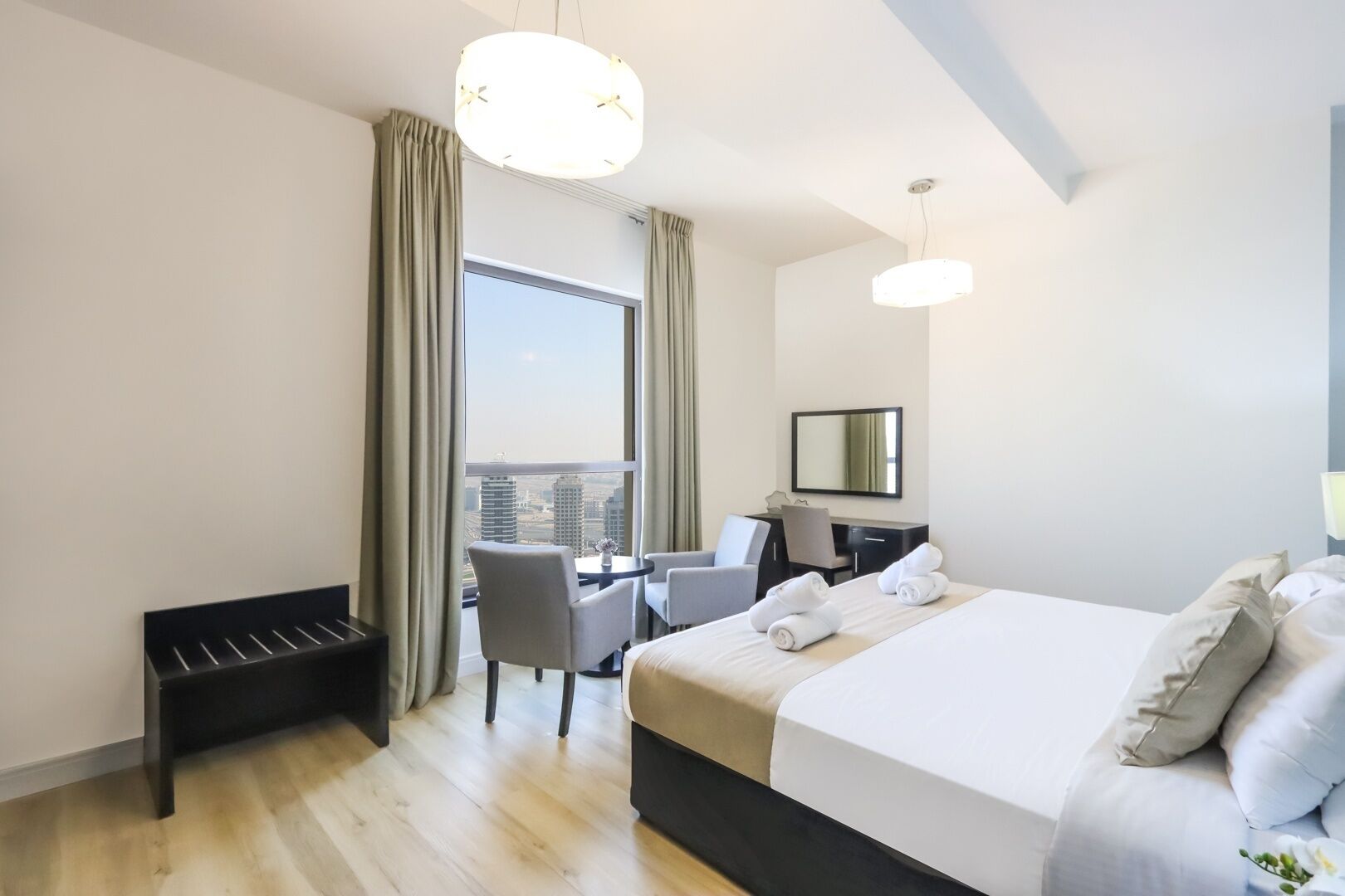 Habitación de hotel con una cama grande, un escritorio, dos sillas, un banco y un ventanal con cortinas.